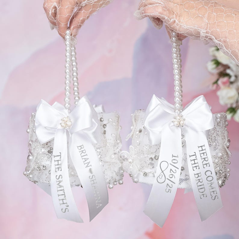 May include: Two white flower baskets featuring pearl-beaded handles, white satin bows, and intricate lace overlays with clear rhinestones. The ribbons are printed with the text THE SMITH'S, BRIAN SHANNA, 10/26/25, and HERE COMES THE BRIDE.