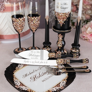 May include: Black and gold wedding cake serving set with a cake knife, two champagne flutes, a cake server, and a candle holder. The set is decorated with rhinestones and the text "Mr. & Mrs. Holsinger 04/22/2023" is engraved on the cake knife and candle holder.