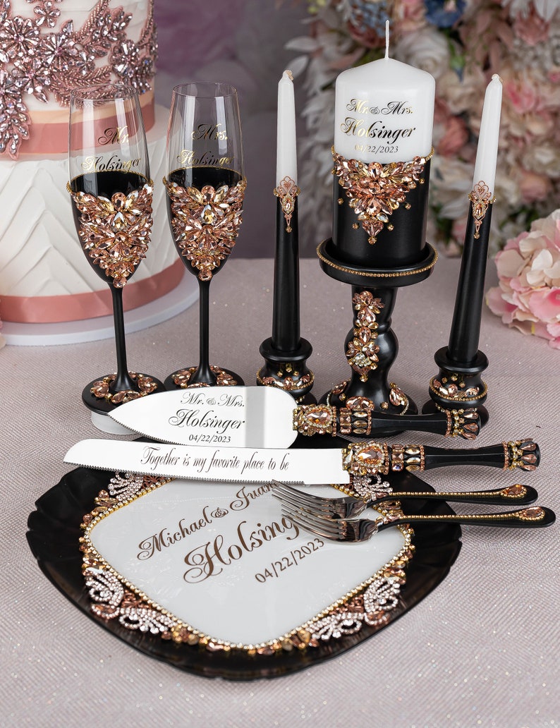 May include: Black and gold wedding cake serving set with a cake knife, two champagne flutes, a cake server, and a candle holder. The set is decorated with rhinestones and the text "Mr. & Mrs. Holsinger 04/22/2023" is engraved on the cake knife and candle holder.