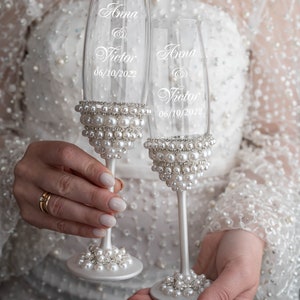 May include: Two champagne flutes decorated with white pearls and the names "Anna & Victor" and the date "06/10/2022" etched on the glass.
