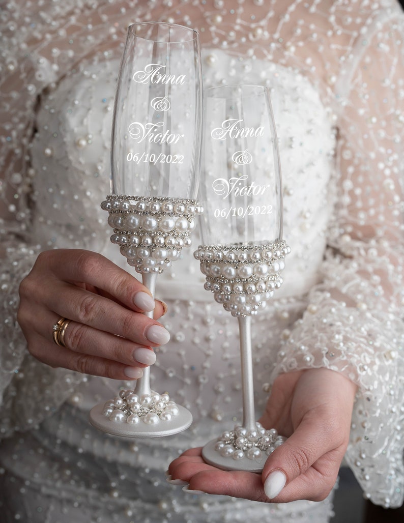 May include: Two champagne flutes decorated with white pearls and the names "Anna & Victor" and the date "06/10/2022" etched on the glass.
