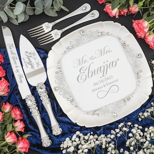 May include: A white cake plate with a silver trim and rhinestones. The plate has the text "Mr. & Mrs. Elnajjar 06/10/2022 Always & Forever" printed on it. A silver cake knife and server with rhinestones and the text "Mr. & Mrs. Elnajjar 06/10/2022 You will Forever be my Always" are on a blue velvet surface.