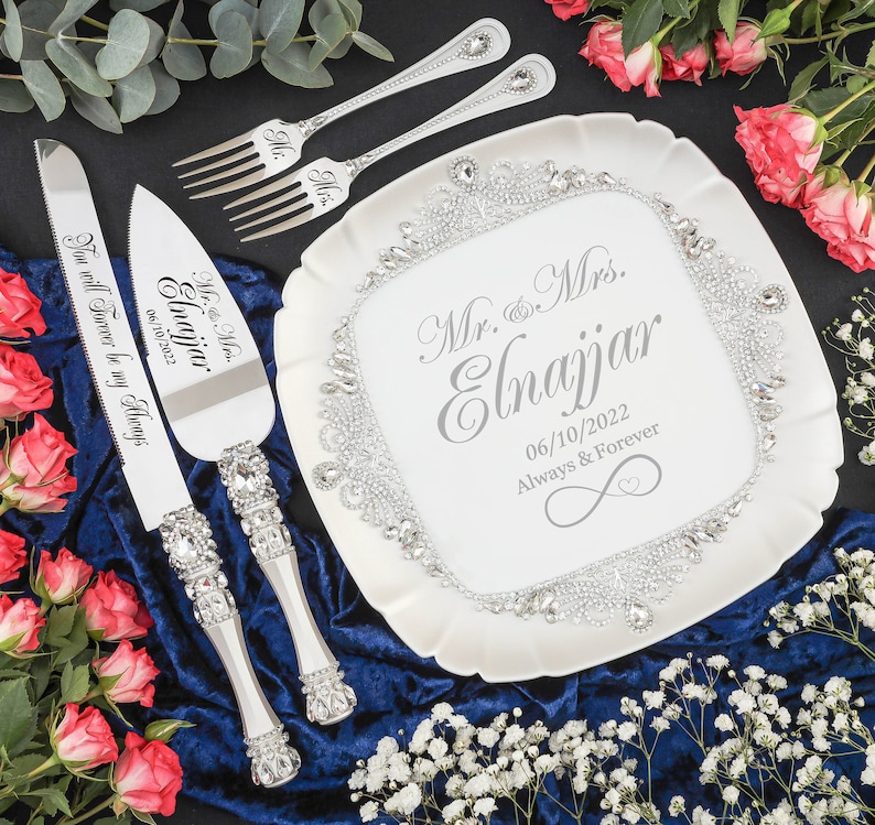 May include: A white cake plate with a silver trim and rhinestones. The plate has the text "Mr. & Mrs. Elnajjar 06/10/2022 Always & Forever" printed on it. A silver cake knife and server with rhinestones and the text "Mr. & Mrs. Elnajjar 06/10/2022 You will Forever be my Always" are on a blue velvet surface.