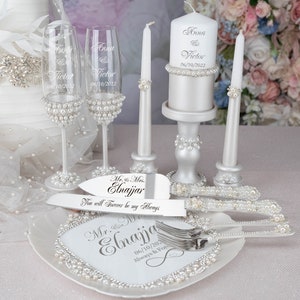 May include: Wedding cake cutting set with a white cake plate, two champagne flutes, a cake knife and server, and two white candles with pearl accents. The cake plate and candles are engraved with the names "Anna & Victor" and the date "06/10/2022". The cake knife and server are engraved with "Mr. & Mrs. Elnajjar" and "You will forever be my always".