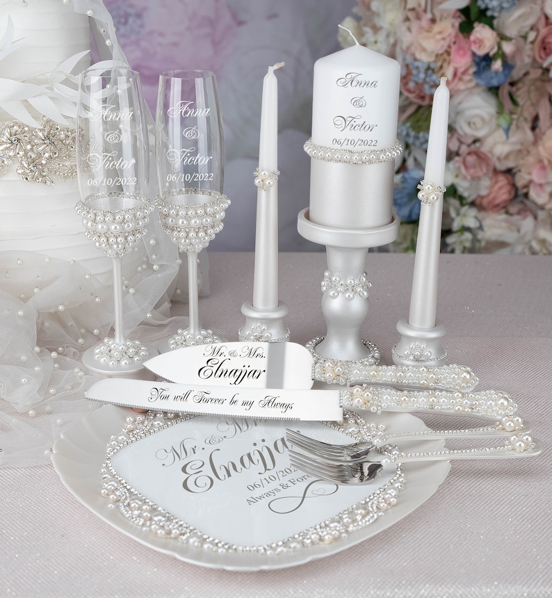 May include: Wedding cake cutting set with a white cake plate, two champagne flutes, a cake knife and server, and two white candles with pearl accents. The cake plate and candles are engraved with the names "Anna & Victor" and the date "06/10/2022". The cake knife and server are engraved with "Mr. & Mrs. Elnajjar" and "You will forever be my always".