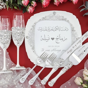 May include: White wedding cake serving set with a white plate, two champagne flutes, a cake knife, and two forks. The plate has a decorative border of rhinestones and the text "دامت أيا حكم عامرة بالمسرات" in Arabic script. The cake knife and forks also have rhinestones and the text "Ramah Nabila" and "You will Forever be my Always" on the handles. The plate also has the text "رمانج - نبيلة" in Arabic script, the date "08/19/2024", and the text "It was Always you". The forks have the text "I do" and "Me too" on the handles.
