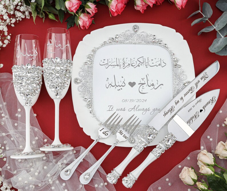 May include: White wedding cake serving set with a white plate, two champagne flutes, a cake knife, and two forks. The plate has a decorative border of rhinestones and the text "دامت أيا حكم عامرة بالمسرات" in Arabic script. The cake knife and forks also have rhinestones and the text "Ramah Nabila" and "You will Forever be my Always" on the handles. The plate also has the text "رمانج - نبيلة" in Arabic script, the date "08/19/2024", and the text "It was Always you". The forks have the text "I do" and "Me too" on the handles.