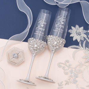 May include: Two clear glass champagne flutes with silver stems, each featuring a large, intricate crystal embellishment at the base of the bowl. The glass is engraved with the names Kejsi and Enea, the date 08.01.2026, and the text Happily Ever After.