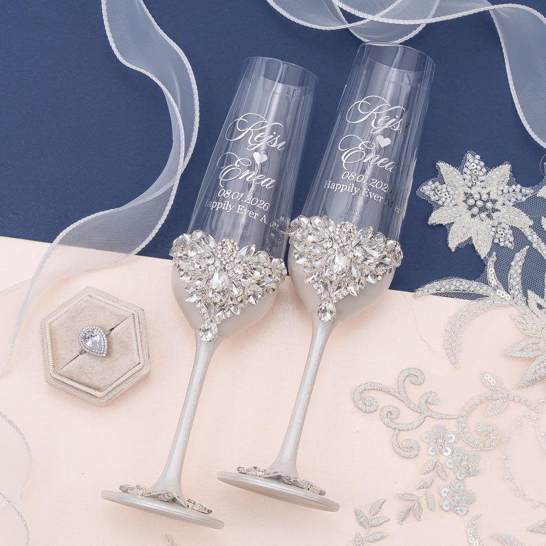 May include: Two clear glass champagne flutes with silver stems, each featuring a large, intricate crystal embellishment at the base of the bowl. The glass is engraved with the names Kejsi and Enea, the date 08.01.2026, and the text Happily Ever After.