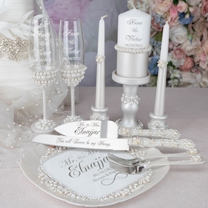 May include: Wedding cake cutting set with a white cake plate, two champagne flutes, a cake knife and server, and two white candles with pearl accents. The cake plate and candles are engraved with the names "Anna & Victor" and the date "06/10/2022". The cake knife and server are engraved with "Mr. & Mrs. Elnajjar" and "You will forever be my always".