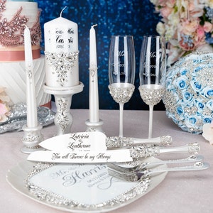 May include: Wedding cake cutting set featuring a white cake knife and server, two champagne flutes, two white candles, and a white cake with silver embellishments. The cake knife and server have a silver handle with rhinestones and the text "Latricia Leathrome - You will Forever be my Always". The champagne flutes have silver bands with rhinestones and the text "Mr. Harris 06/15/2023". The candles are white with silver bands and rhinestones. The cake is white with silver embellishments and the text "Mavis 05/15/13".
