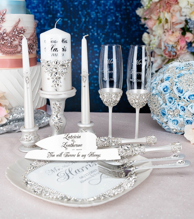 May include: Wedding cake cutting set featuring a white cake knife and server, two champagne flutes, two white candles, and a white cake with silver embellishments. The cake knife and server have a silver handle with rhinestones and the text "Latricia Leathrome - You will Forever be my Always". The champagne flutes have silver bands with rhinestones and the text "Mr. Harris 06/15/2023". The candles are white with silver bands and rhinestones. The cake is white with silver embellishments and the text "Mavis 05/15/13".