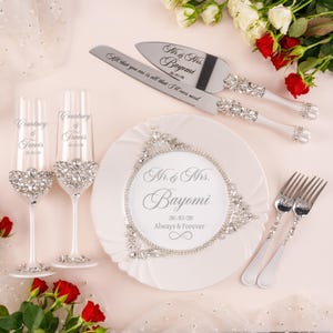 May include: A wedding set featuring two champagne flutes, a cake knife, a cake server, and two forks. The items are decorated with clear crystals. The flutes are engraved with Courtney & Travis 26/10/26. The plate reads Mr. & Mrs. Bayomi 26/10/26 Always & Forever.