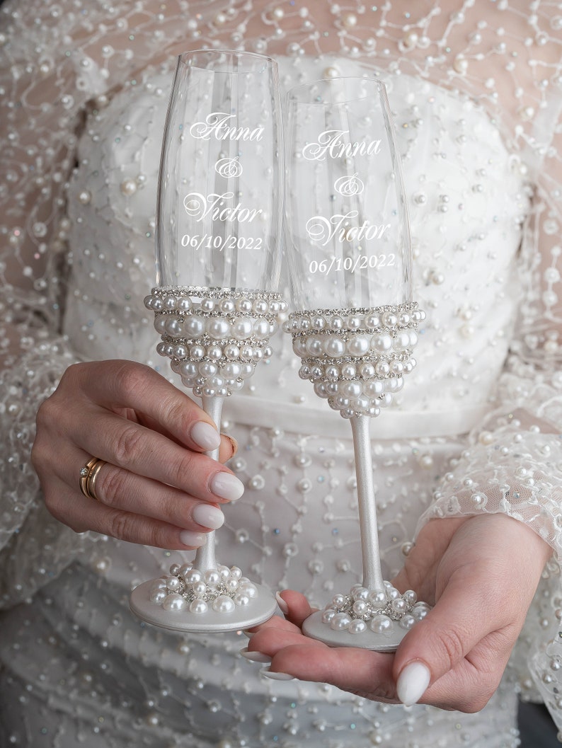 May include: Two champagne flutes with pearl embellishments. The flutes are engraved with the names "Anna & Victor" and the date "06/10/2022".