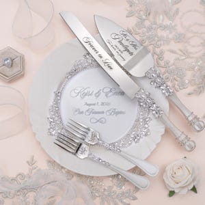 May include: A silver cake knife and server set with crystal-encrusted handles, paired with two matching forks. The knife is engraved with "Forever in Love" and the server with "Mr. & Mrs. Pauligoti". A white plate features "Kejsi & Enea" and "August 1, 2026".