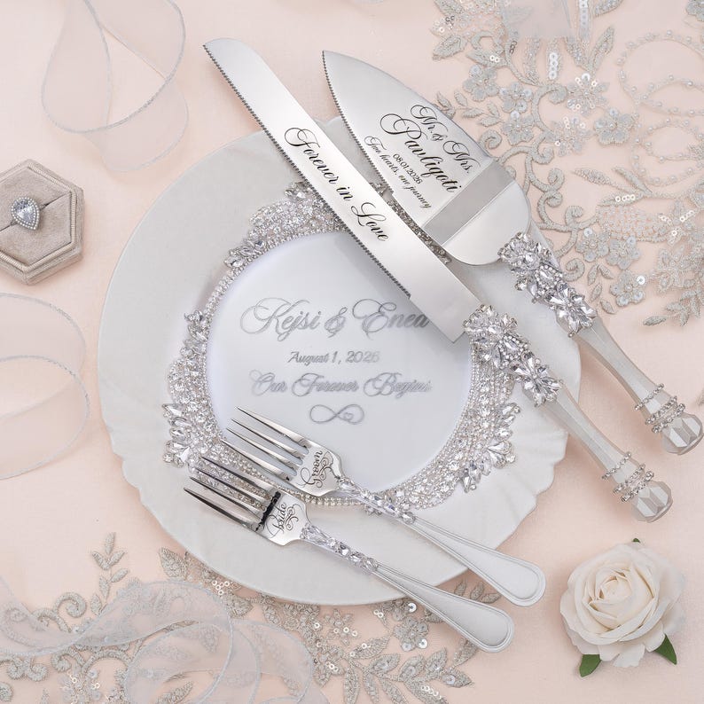 May include: A silver cake knife and server set with crystal-encrusted handles, paired with two matching forks. The knife is engraved with "Forever in Love" and the server with "Mr. & Mrs. Pauligoti". A white plate features "Kejsi & Enea" and "August 1, 2026".
