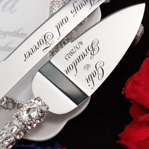 May include: A silver cake knife and server set with a rhinestone handle. The knife has the text "Always and Forever" engraved on it. The server has the text "Gary & Brianna 9/13/2023" engraved on it.