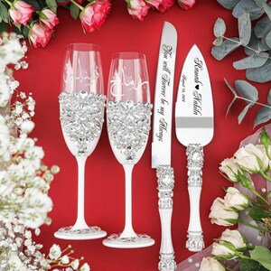 May include: A white wedding cake serving set with two champagne flutes and a cake knife. The flutes and knife are decorated with rhinestones. The cake knife has the text "Ramah Nabila August 18, 2023" and "You will forever be my Always" engraved on it.