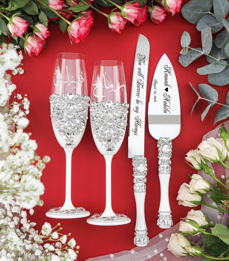May include: A white wedding cake serving set with two champagne flutes and a cake knife. The flutes and knife are decorated with rhinestones. The cake knife has the text "Ramah Nabila August 18, 2023" and "You will forever be my Always" engraved on it.