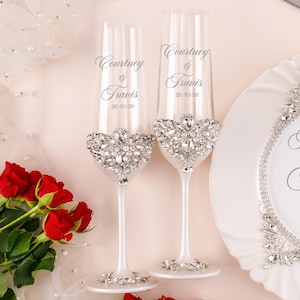 May include: Two clear glass champagne flutes with white stems, each featuring a cluster of clear rhinestones at the base of the bowl and on the foot. The glasses are engraved with the names Courtney & Travis and the date 26/10/26 in script.
