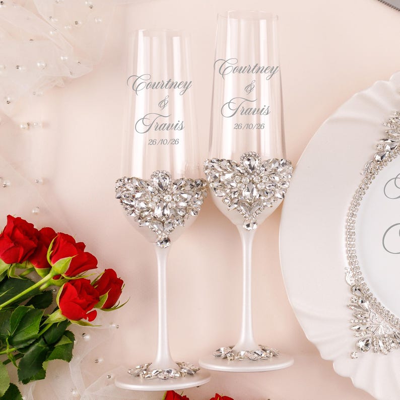 May include: Two clear glass champagne flutes with white stems, each featuring a cluster of clear rhinestones at the base of the bowl and on the foot. The glasses are engraved with the names Courtney & Travis and the date 26/10/26 in script.