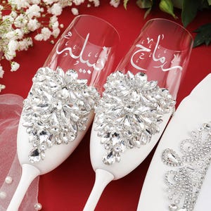 May include: Two white champagne flutes with a decorative crystal design. The flutes are engraved with Arabic calligraphy.