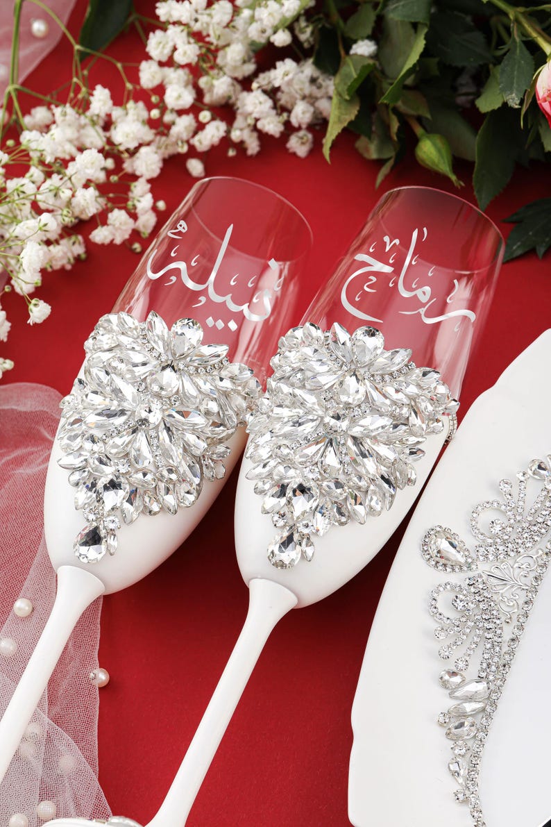May include: Two white champagne flutes with a decorative crystal design. The flutes are engraved with Arabic calligraphy.