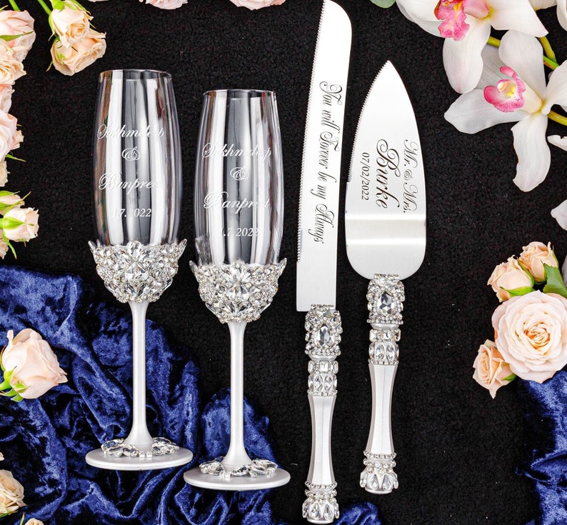 May include: A wedding cake cutting set with two champagne flutes and a cake knife. The flutes and knife are decorated with rhinestones. The flutes are engraved with the names "Khmer" and "Banpre" and the date "07. 2022". The cake knife is engraved with "Mr. & Mrs. Burke" and the date "07/02/2022". The knife also has the text "You will Forever be my Always".
