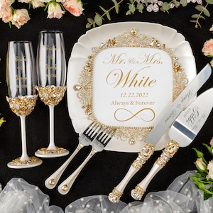 May include: A white cake plate with gold trim and rhinestones, a gold infinity symbol with a heart, and the text "Mr. & Mrs. White 12.22.2022 Always & Forever". Two champagne flutes with gold and rhinestone trim, a cake knife and server with gold and rhinestone handles, and a fork with a gold handle and the text "I do" are also included.