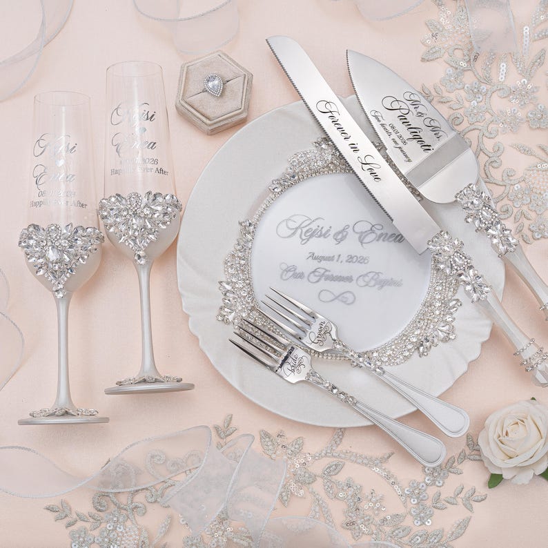 May include: Two champagne flutes, a cake knife, a cake server, and two forks, all featuring silver-toned metal and clear crystal embellishments. A white plate with the text Kejsi & Enea August 1, 2026 Our Forever Begins and a ring box are included.