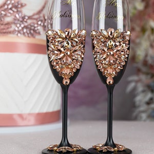 May include: Two black champagne flutes with gold lettering that reads "Mr. Holsinger" and "Mrs. Holsinger". The flutes are decorated with a band of rose gold rhinestones.