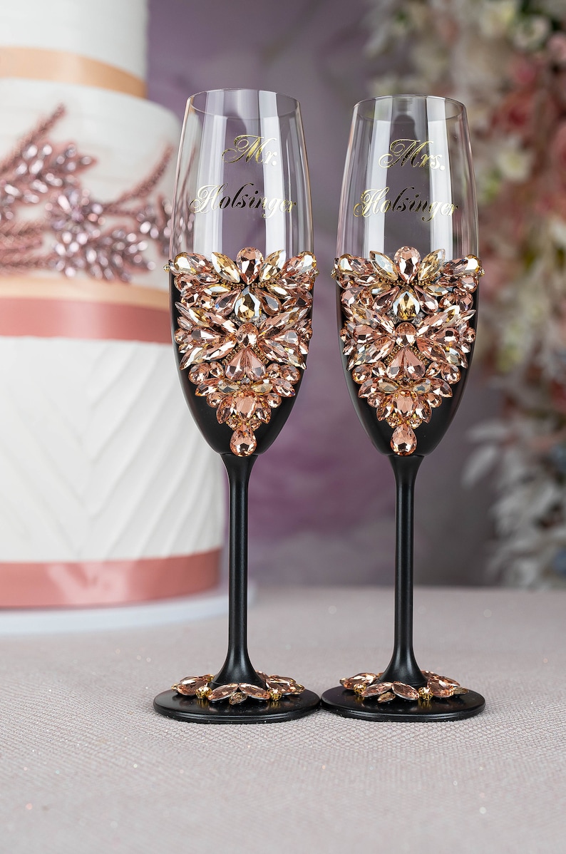 May include: Two black champagne flutes with gold lettering that reads "Mr. Holsinger" and "Mrs. Holsinger". The flutes are decorated with a band of rose gold rhinestones.