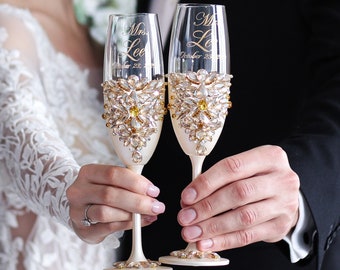 May include: Two champagne flutes with cream-colored stems and bases. The clear glass flutes are adorned with gold and crystal embellishments. The flutes are engraved with the names "Mrs. Lee" and the date "October 23, 2021".
