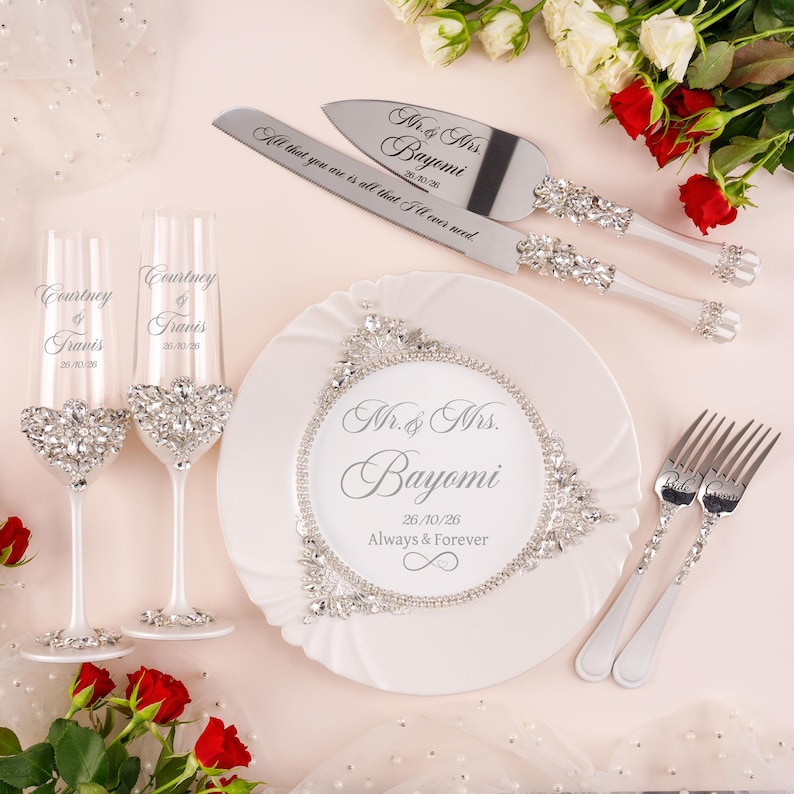 May include: A wedding set featuring two champagne flutes, a cake knife, a cake server, and two forks. The items are decorated with clear crystals. The flutes are engraved with Courtney & Travis 26/10/26. The plate reads Mr. & Mrs. Bayomi 26/10/26 Always & Forever.
