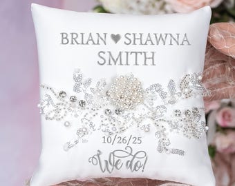 Peut inclure: Un coussin d'alliances carré en satin blanc avec le texte imprimé BRIAN SHAWNA SMITH, la date 10/26/25 et la phrase We do!. Le centre est orné d'une application florale argentée agrémentée de strass transparents et de perles blanches.