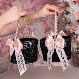 May include: Two black and sequined flower girl baskets with pearl handles and pink satin bows. One basket has a ribbon with the text "Love Never Fades Away" and the other has a ribbon with the text "Toni Juan July 20 2023".