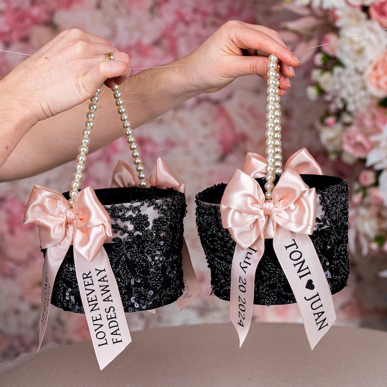 May include: Two black and sequined flower girl baskets with pearl handles and pink satin bows. One basket has a ribbon with the text "Love Never Fades Away" and the other has a ribbon with the text "Toni Juan July 20 2023".