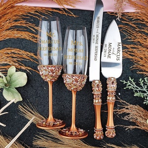May include: A wedding cake cutting set with two champagne flutes and a knife. The flutes and knife are decorated with rhinestones and have the names "Marcus" and "Monae" engraved on them, along with the date "08 26 23".