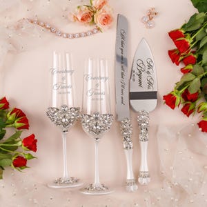 May include: Two clear glass champagne flutes, a cake knife, and a cake server, all featuring white handles and stems adorned with clear crystals. The flutes are engraved with Courtney & Travis 26/10/26, the knife with All that you are is all that I'll ever need, and the server with Mr. & Mrs. Bayomi 26/10/26.