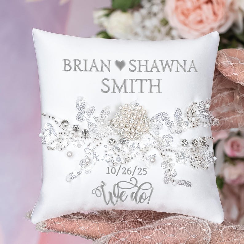 May include: White satin pillow with a silver embroidered design, featuring the names "BRIAN SHAWNA SMITH" and the date "10/26/25" with the text "We do!"