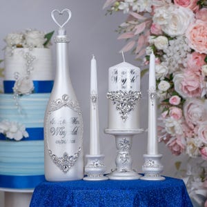 May include: A white bottle with a heart-shaped topper, two thin taper candles, and one thick pillar candle on a pedestal. The items feature silver script text reading Mr. & Mrs. Winfield 09/26/2026 and are decorated with clear crystal embellishments.