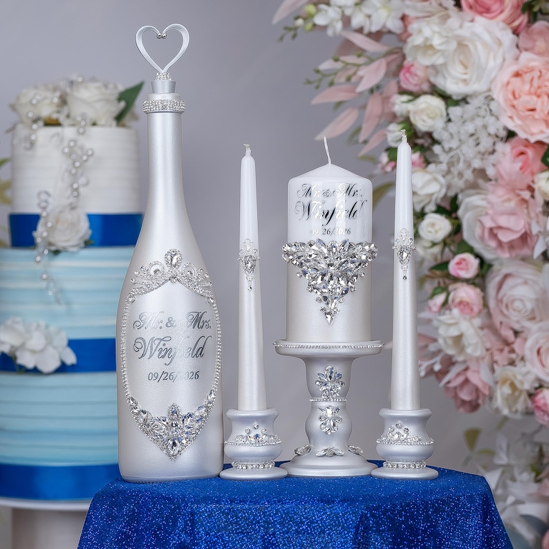 May include: A white bottle with a heart-shaped topper, two thin taper candles, and one thick pillar candle on a pedestal. The items feature silver script text reading Mr. & Mrs. Winfield 09/26/2026 and are decorated with clear crystal embellishments.