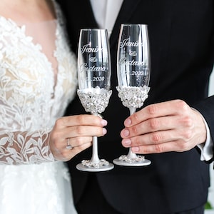 May include: Two champagne flutes with silver rims and rhinestones around the base. The flutes are engraved with the names "Janine & Gustavo" and the date "07.2020".