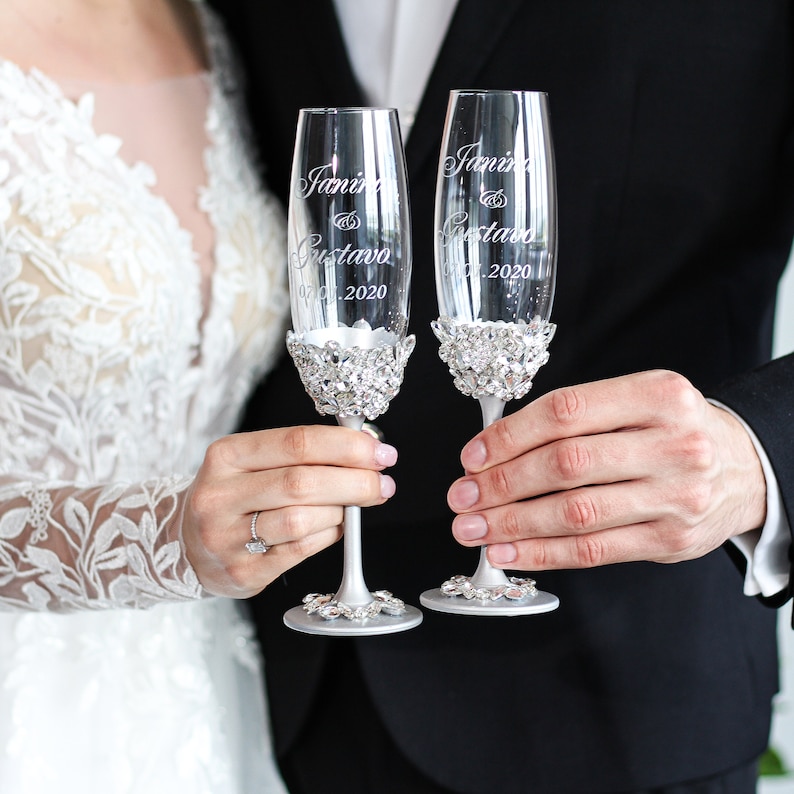 May include: Two champagne flutes with silver rims and rhinestones around the base. The flutes are engraved with the names "Janine & Gustavo" and the date "07.2020".