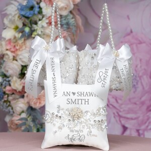 May include: Three white satin pillow bags with pearl handles and silver ribbon. The bags are decorated with lace and rhinestones. The ribbons are personalised with the names "Brian & Shawna", "The Smith's", and "Here Comes The Bride". The date "10/26/25" is printed on two of the ribbons. A white satin pillow with a silver heart and the text "An Shawni Smith" is in the centre of the image. The pillow is decorated with rhinestones and lace. The date "10/26/25" and the text "We do!" are printed on the pillow.