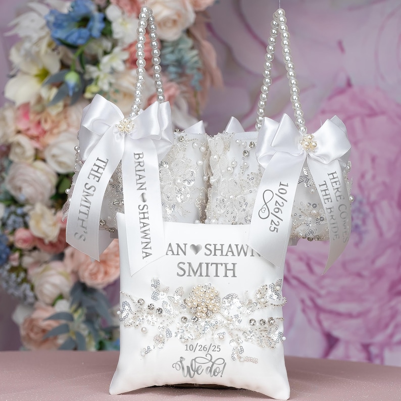 May include: Three white satin pillow bags with pearl handles and silver ribbon. The bags are decorated with lace and rhinestones. The ribbons are personalised with the names "Brian & Shawna", "The Smith's", and "Here Comes The Bride". The date "10/26/25" is printed on two of the ribbons. A white satin pillow with a silver heart and the text "An Shawni Smith" is in the centre of the image. The pillow is decorated with rhinestones and lace. The date "10/26/25" and the text "We do!" are printed on the pillow.