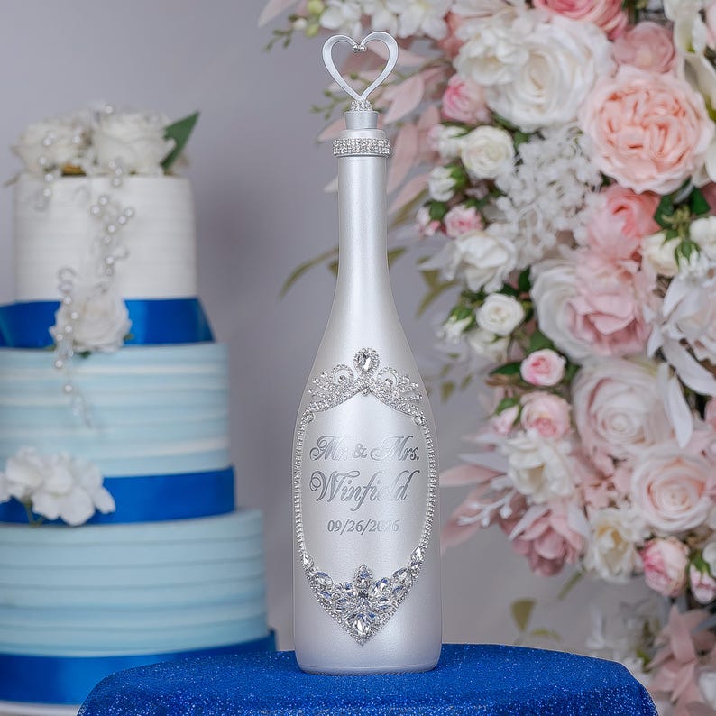 May include: A matte white bottle featuring a silver heart-shaped topper, a rhinestone-encrusted neck, and a central oval frame of clear crystals. The bottle is engraved with the text "Mr. & Mrs. Winfield" and the date "09/26/2026" in script font.