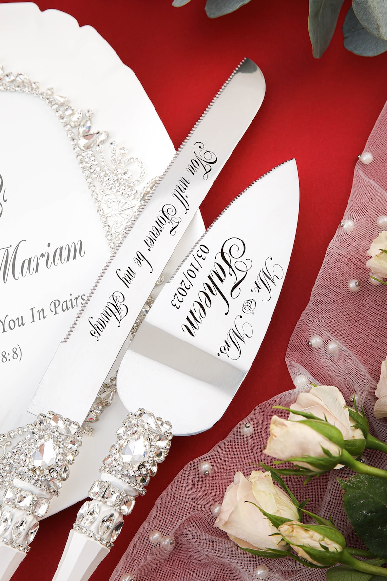 May include: A white cake server set with rhinestone handles. The server blades are engraved with the text "Mr. & Mrs. Faheem 03/10/2023" and "Forever be my Always Mariam You In Pairs 8:8)".