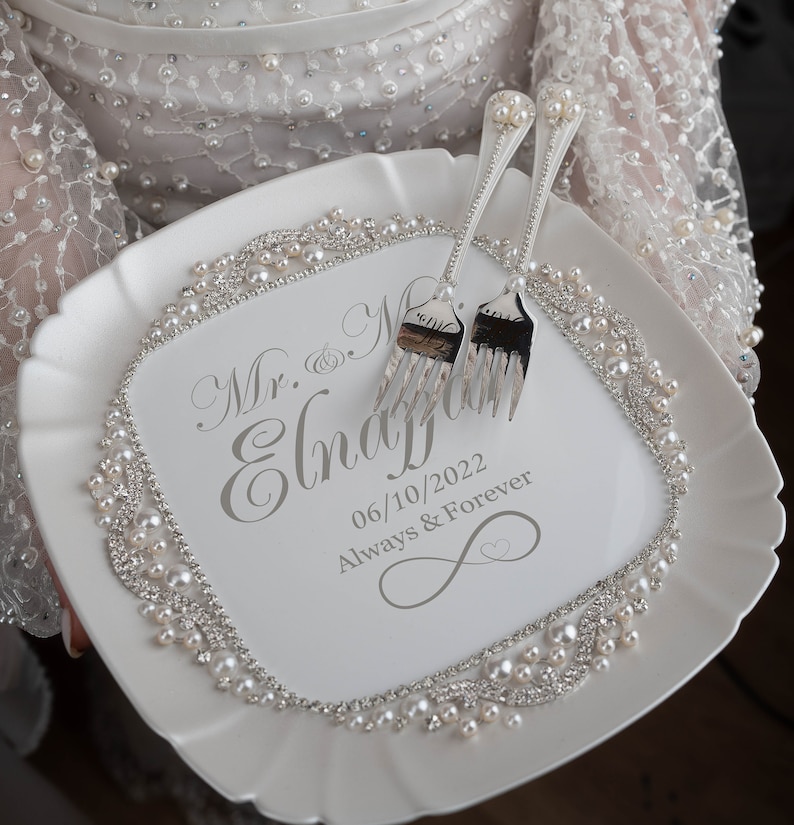 May include: White ceramic cake plate with a pearl and rhinestone border. The plate is engraved with the text "Mr. & Mrs.  Engaged 06/10/2022 Always & Forever" and features a silver infinity symbol. Two silver forks are placed on the plate.