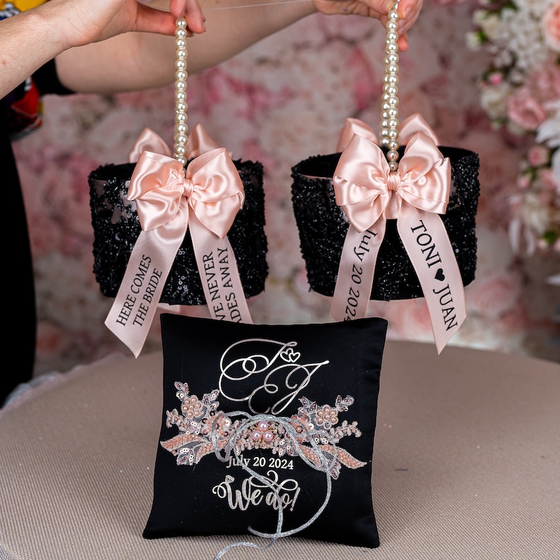 May include: Two black and white sequined ring bearer pillows with pink satin bows and personalized ribbons. The ribbons say "Here comes the bride", "We never fades away", and "Toni Juan July 20 2024". A black satin pillow with a silver embroidered design and the text "July 20 2024 We do!" is in the center.